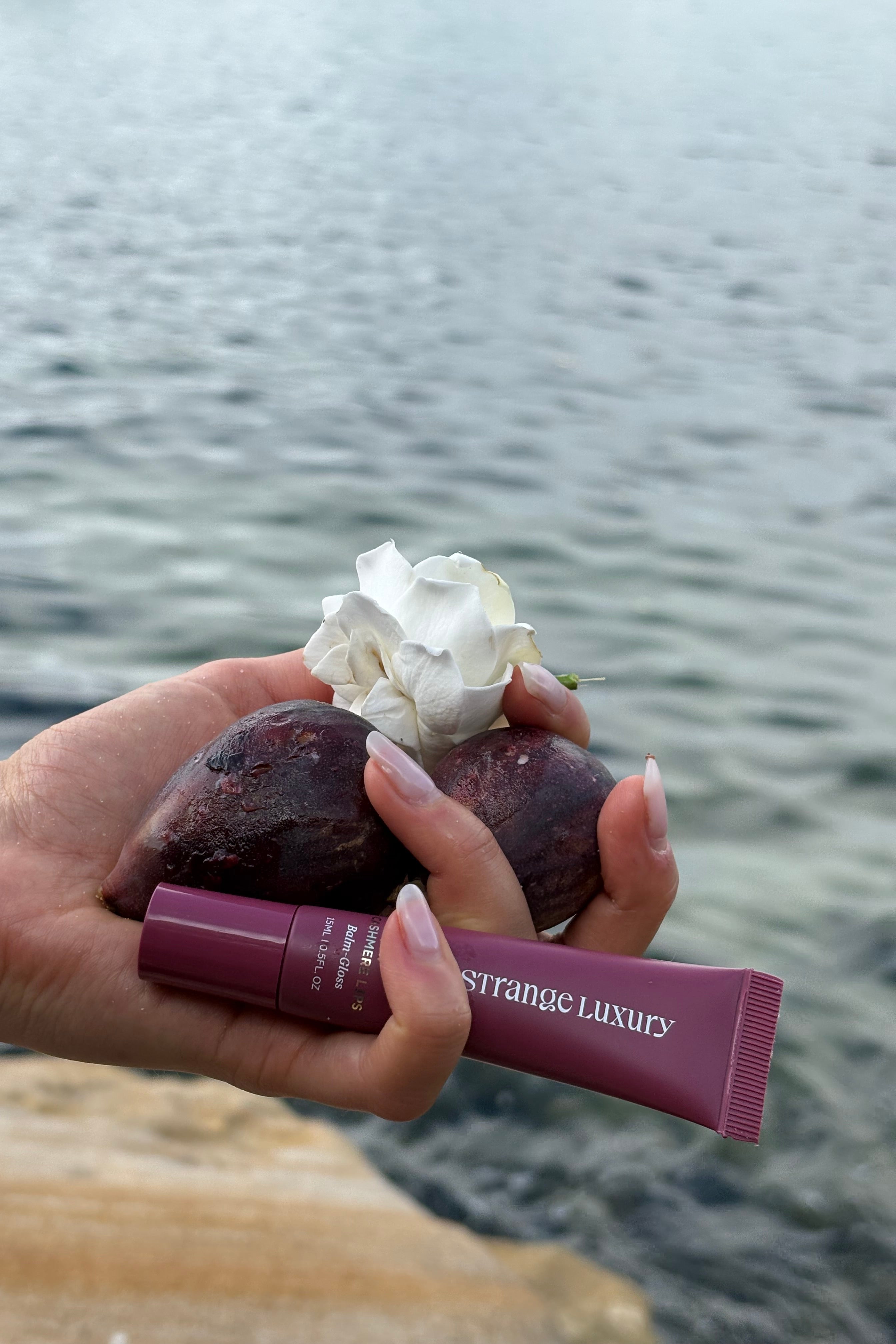 Hand holding a purple tube of Strange Luxury product with figd and flowers against the ocean. 