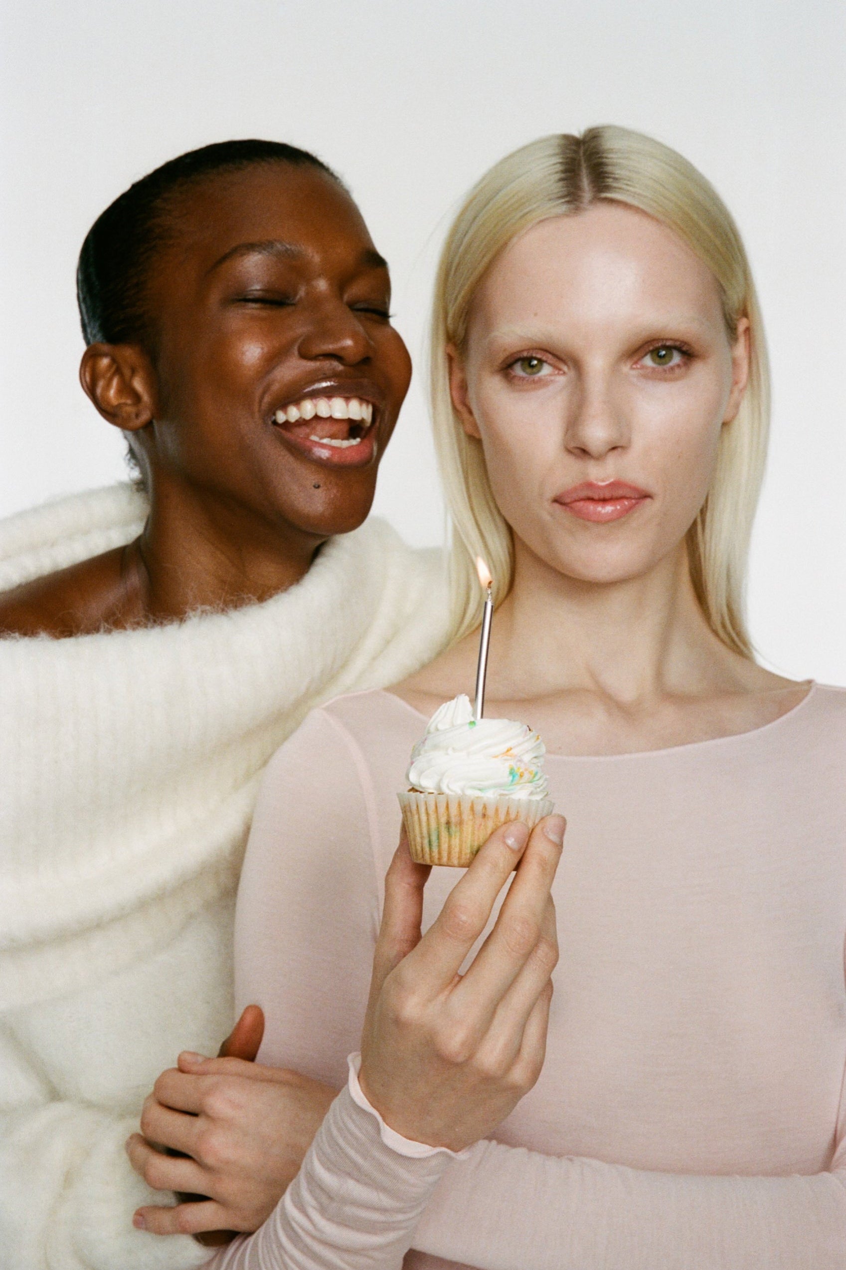 Two women, one holding a cupcake with a lit candle, against a white background.
