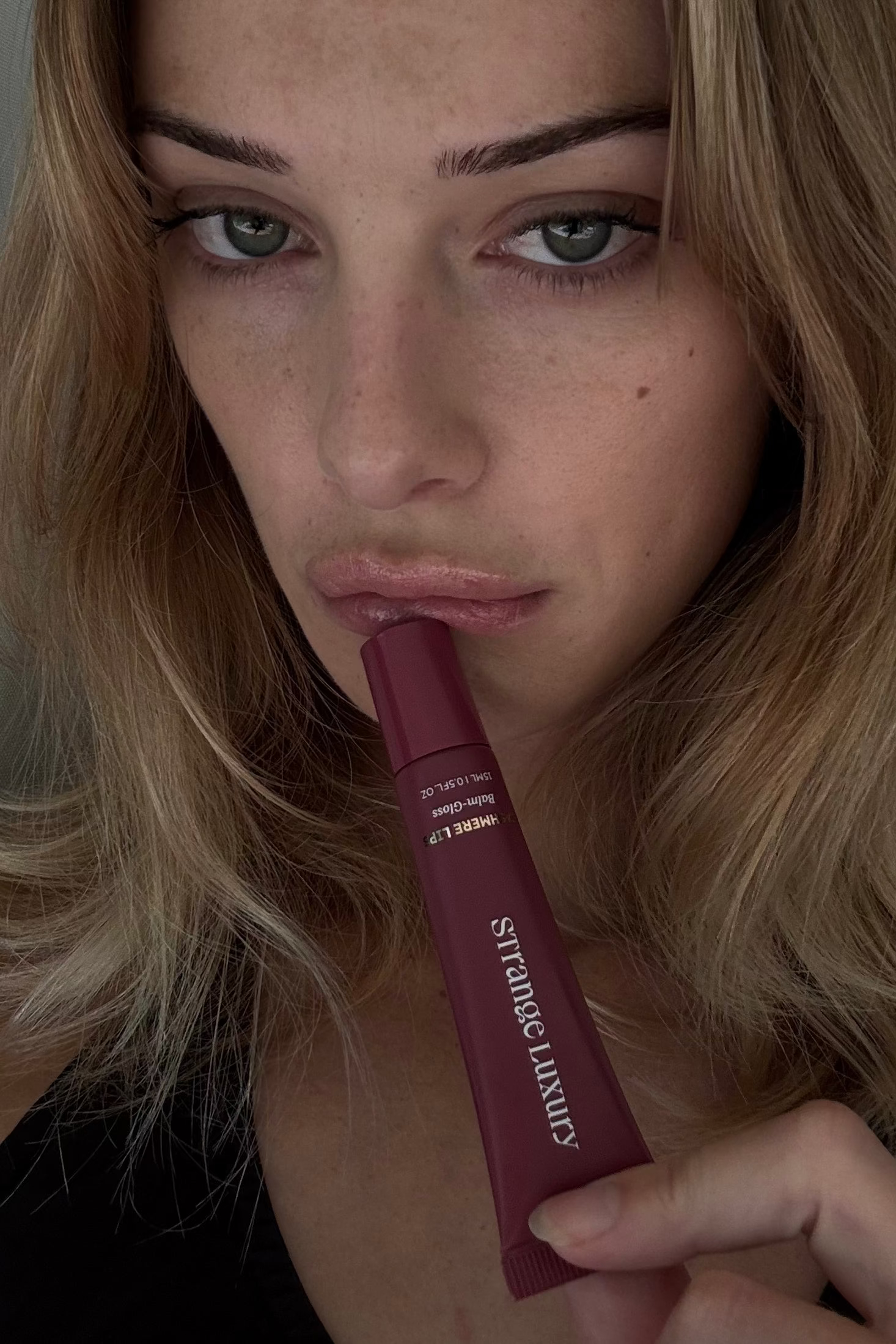 Woman holding a Cherry Cashmere lip gloss with 'Strange Luxury' branding.