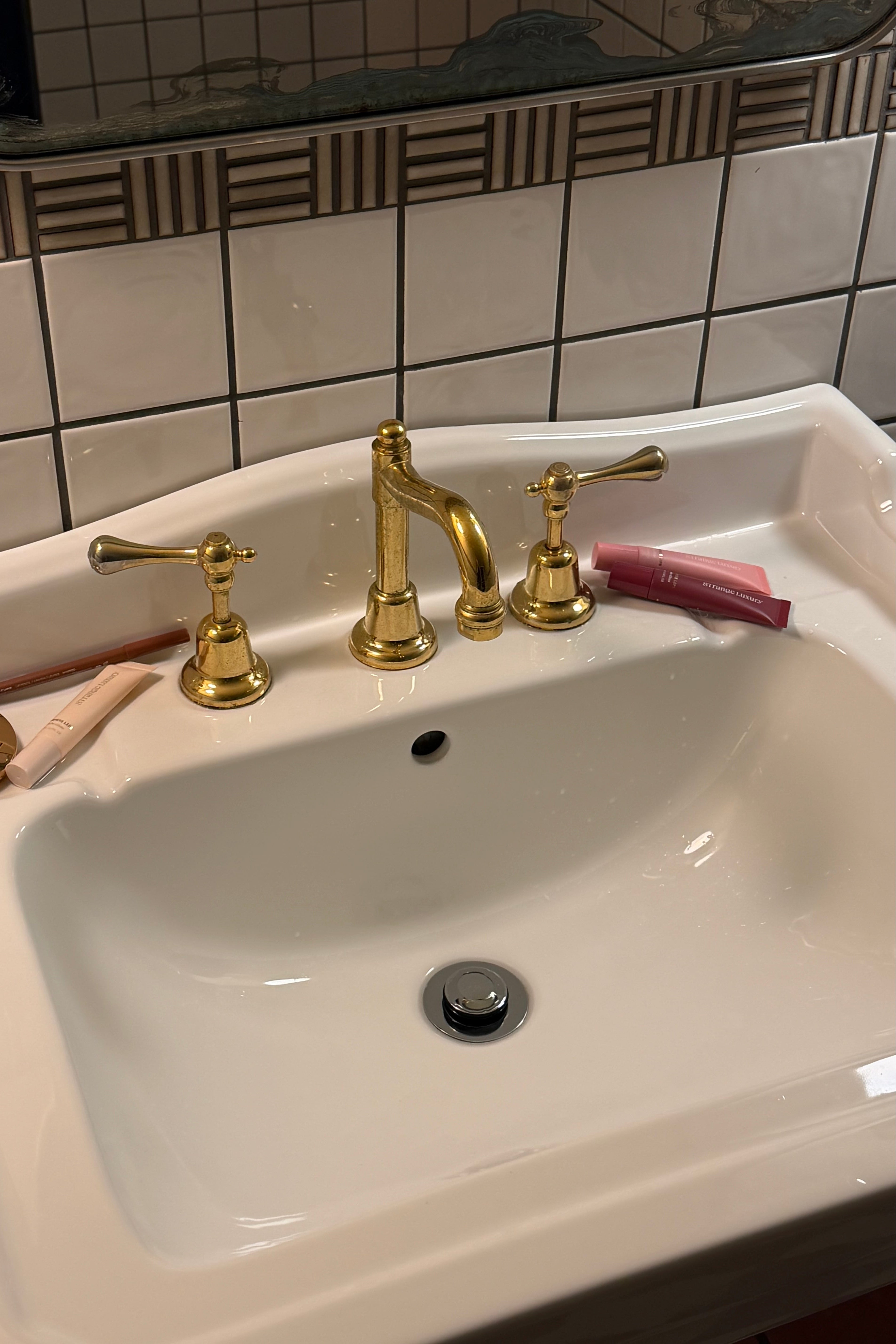White sink with gold faucet and handles on a tiled bathroom wall with lip products.